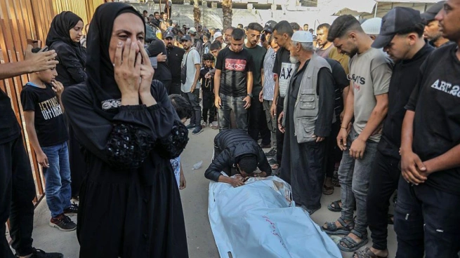 A man kneels to kiss a body bag on the ground as others gather, while a woman weeps with her face covered.